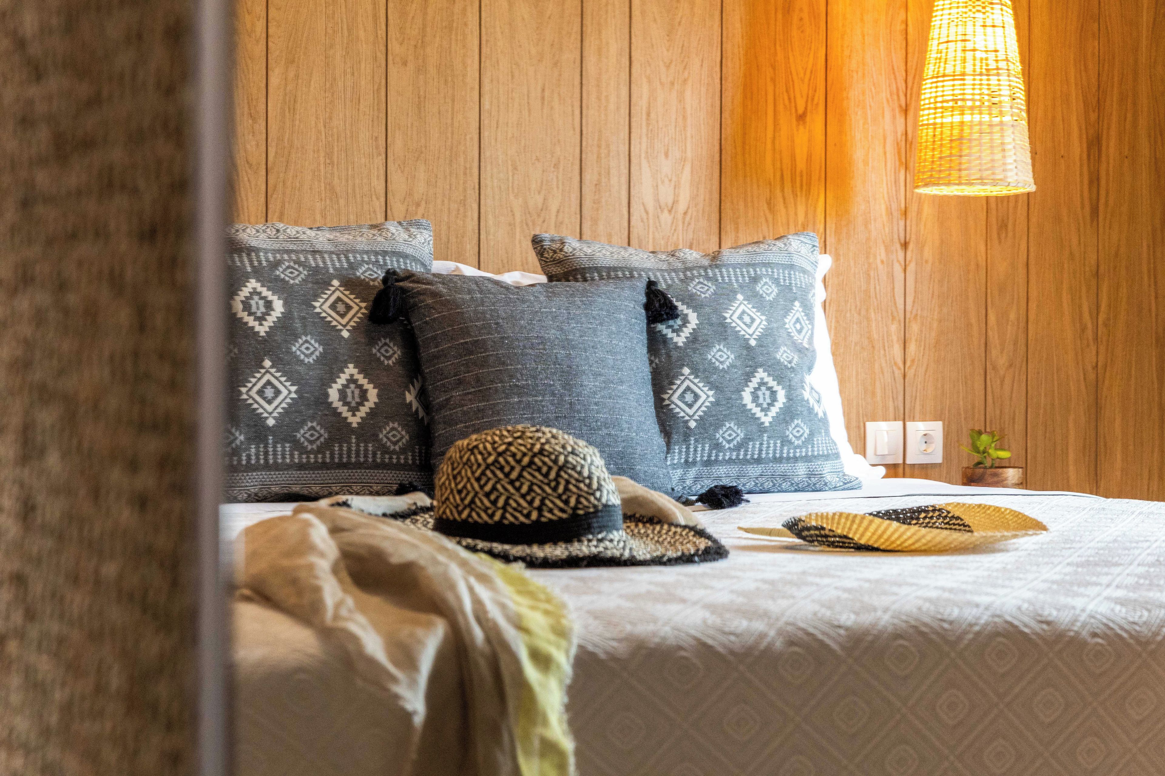 A neatly made bed with patterned pillows, a woven hat, and a small plant on a wooden wall.