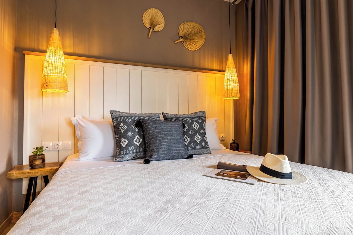 A neatly made bed with patterned pillows, two wall-mounted lamps, and a hat on the bed.