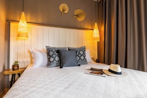 Un lit bien fait, agrémenté d’oreillers à motifs, deux lampes fixées au mur et un chapeau posé sur le lit.