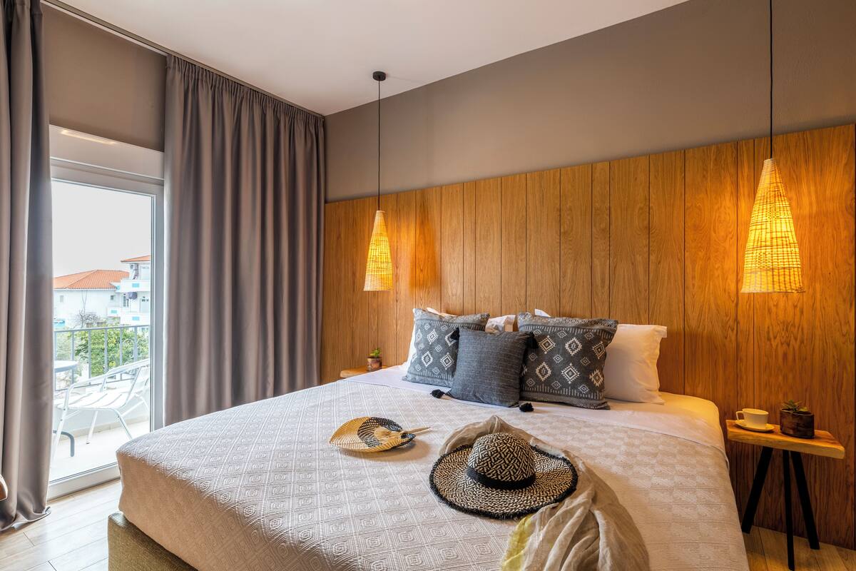 A modern hotel room with a large bed, two hanging pendant lights, a view of the outdoors, and a small table with a cup and a plant.