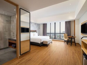 A hotel room with a large bed, a desk, a chair, a TV, and a view of the city. - Luminor Hotel Padjadjaran Bogor (Bogor)