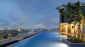 Una piscina infinita en la azotea con vistas a la ciudad, un edificio moderno con iluminación cálida y un cielo despejado.