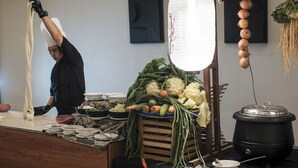 Una cocina con una persona preparando fideos, una variedad de verduras frescas y una olla en la encimera.