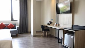 Habitación de hotel con un sofá gris, un escritorio pequeño con una silla, un televisor montado en la pared y un refrigerador pequeño.