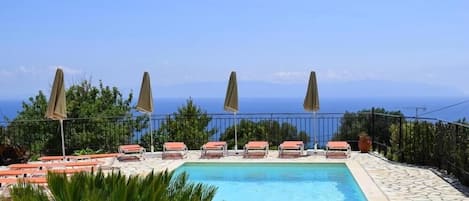 A swimming pool with lounge chairs and umbrellas, surrounded by a stone deck and a metal fence, with a view of the sea and mountains in the background.