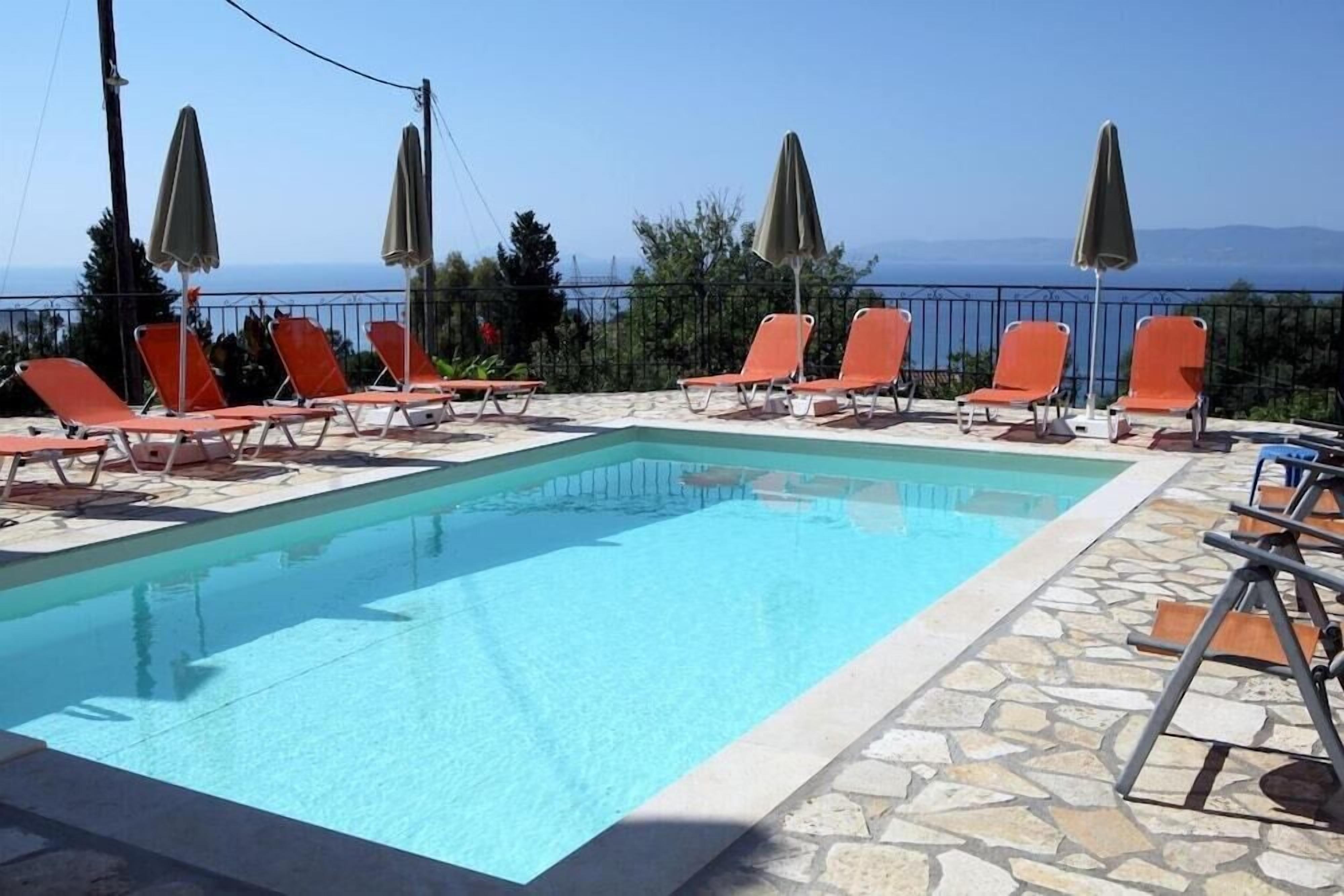 En rektangulär swimmingpool med kristallklart blått vatten, omgiven av orange solstolar och gröna parasoller, med en bakgrund av träd och en avlägsen utsikt över havet.