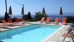 A rectangular swimming pool with clear blue water, surrounded by orange lounge chairs and green umbrellas, set against a backdrop of trees and a distant view of the sea.
