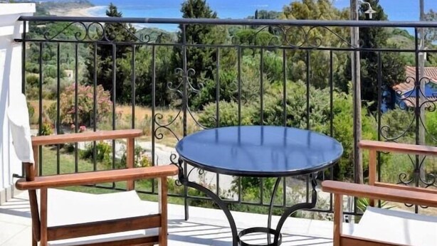 A balcony with a round table and two wooden chairs, overlooking a coastal landscape with trees and a view of the sea.