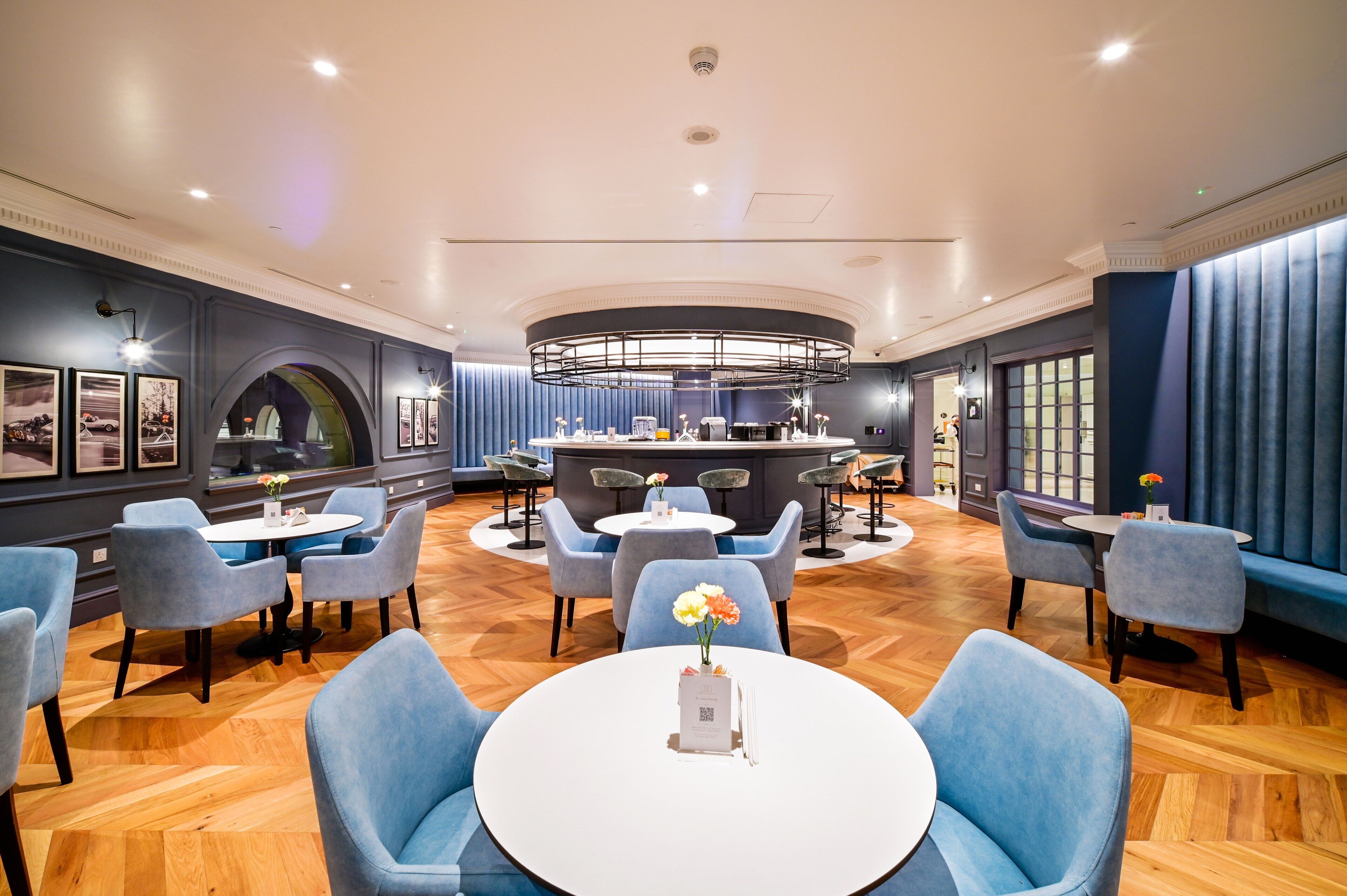 A modern hotel lobby with round tables, blue upholstered chairs, and a central bar area.