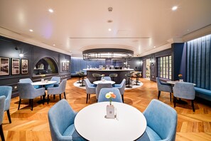 A modern hotel lobby with round tables, blue upholstered chairs, and a central bar area. - 21 High Street Residence (Doha)