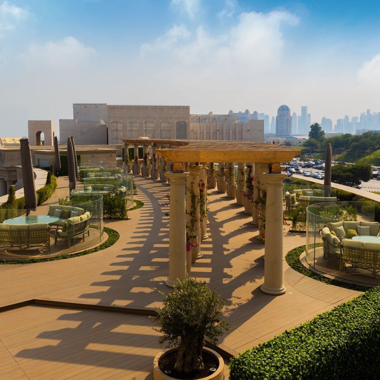 An outdoor seating area with round tables and chairs, surrounded by columns with climbing plants, under a clear sky.