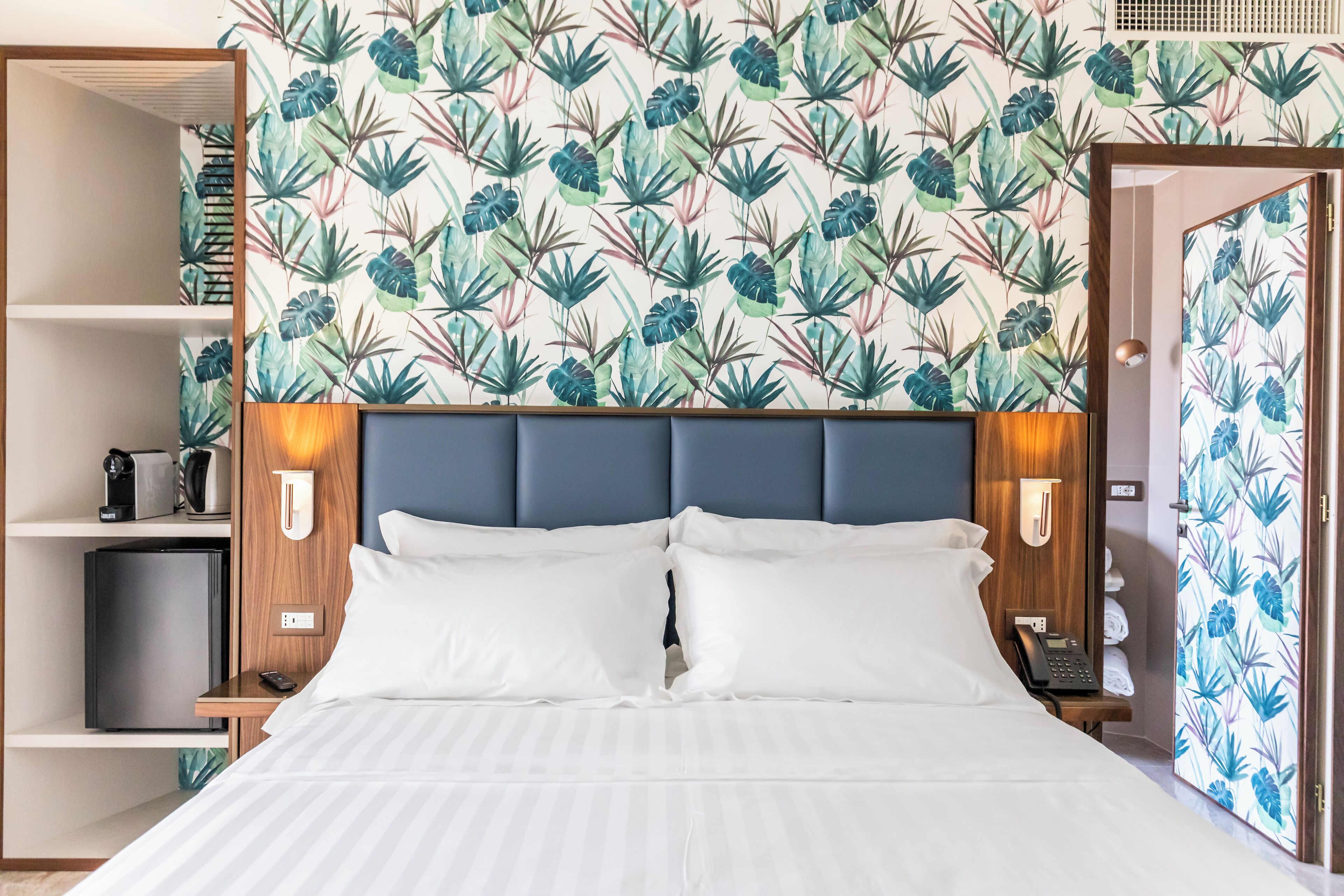 A hotel room with a bed, a headboard, a nightstand, a telephone, and a shelf with a coffee maker.
