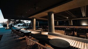 Eine Außenbar mit Holztischen und -stühlen, großen Säulen und Blick auf das Meer.