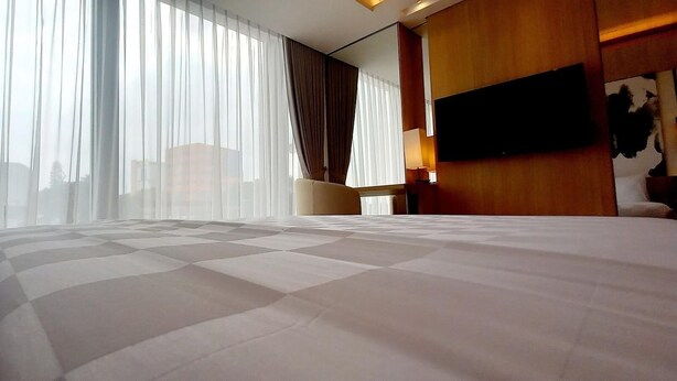 A hotel room with a large bed, a flat-screen TV mounted on the wall, and a window with sheer curtains. - LK Hotel Chadstone Cikarang (Cikarang)