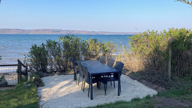 Een tafel met stoelen op een zandstrand, met uitzicht op de zee en heuvels in de verte.