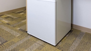 Ein weißer Kühlschrank steht in einer Ecke des Raumes, der einen gemusterten Teppichboden hat.