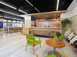 Una cafetería moderna con piso de baldosas, mesas y sillas de madera, una barra con televisor y plantas en macetas.