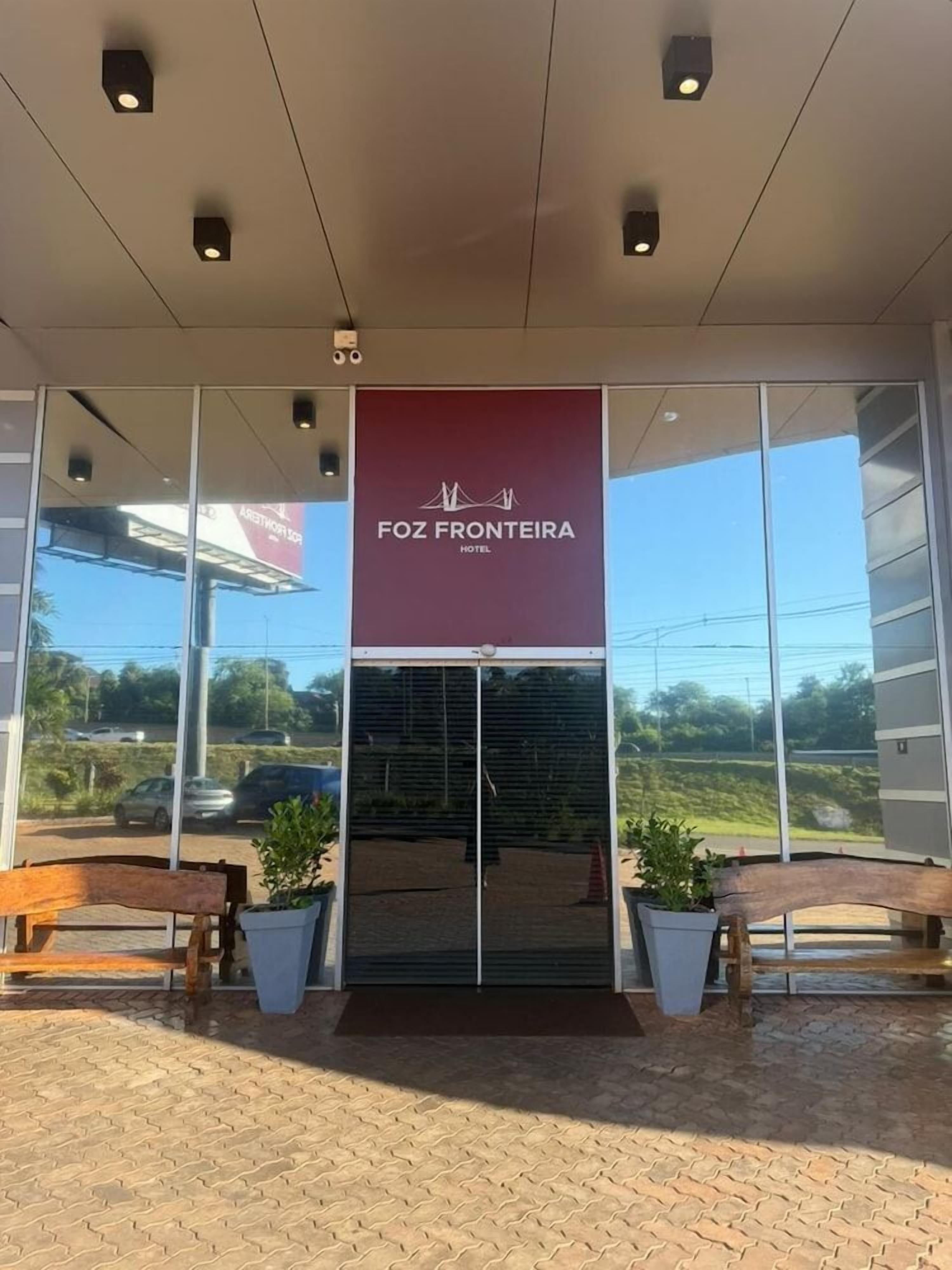 Entrada do hotel Foz Fonteria, com vista livre para a área de estacionamento e um banco na parte externa.