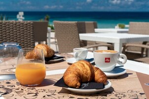 Un área de comedor al aire libre con croissants, un vaso de jugo de naranja y una taza de café, con vista a una playa.