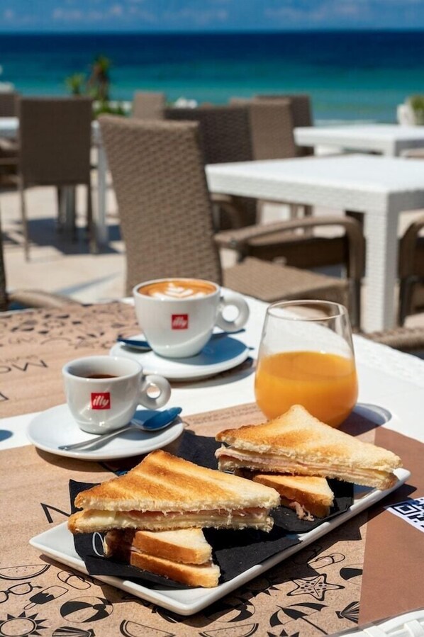 Una mesa con un sándwich, una taza de café y un vaso de zumo de naranja en una mesa frente a la playa.