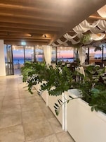 Un restaurante con vista al atardecer sobre el agua, con una decoración interior moderna en tonos blancos y plantas en macetas.