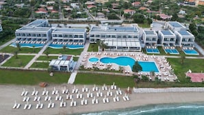 An aerial view of a resort with multiple buildings, a large central pool, and a beach area with sun loungers. - Sea Coast Resort (Polygyros)