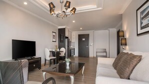 A modern living room with a flat-screen TV, a dining area with a table and chairs, a kitchen area with cabinets, and a sofa with a decorative pillow. - Sirin Exclusive Hotel and Residence (Pattaya)