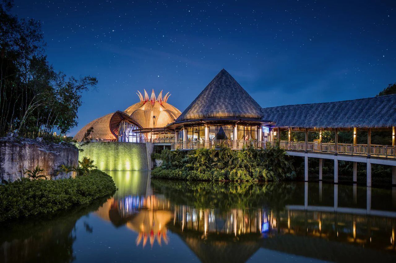 Une vue nocturne d’un complexe hôtelier composé de deux bâtiments distincts : l’un est surmonté d’un dôme, l’autre présente une structure en forme de pyramide, et les deux sont reliés par un pont qui enjambe une étendue d’eau calme.
