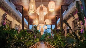 A wooden pathway flanked by lush tropical plants, leading to a thatched-roof building with hanging lanterns. - Jungala Hotel at Vidantaworld Riviera Maya (Playa del Carmen)