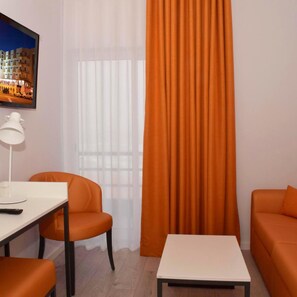 Une pièce avec un bureau blanc, deux chaises orange, une table blanche et un grand rideau orange.
