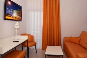 A modern hotel room with a flat-screen TV mounted on the wall, a white desk with a lamp, an orange sofa, and a small table. - THE FOZ Beach Hotel (Figueira da Foz)