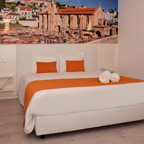 Une chambre avec un grand lit, une literie blanche et des coussins orange. Un grand tableau mural représentant un paysage urbain orne le mur derrière le lit.