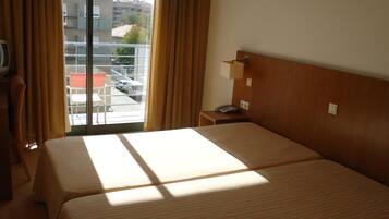 A hotel room with two beds, a window with curtains, a bedside table with a lamp, and a view of the outside.