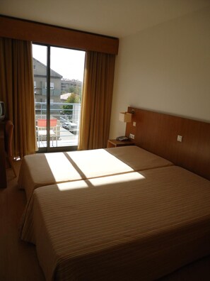 A hotel room with two beds, a window with curtains, a bedside table with a lamp, and a view of the outside. - Hotel Jose Alberto (Viseu)