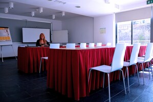 Un salón de conferencias con una larga mesa, sillas blancas, una pizarra y una pantalla para presentaciones.