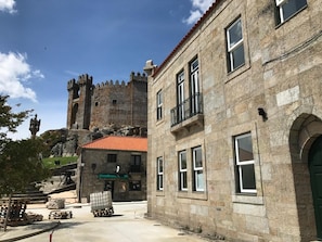 A stone building with a red-tiled roof, a balcony, and a view of a castle-like structure in the background. - Penedono Medieval Hotel (Penedono)
