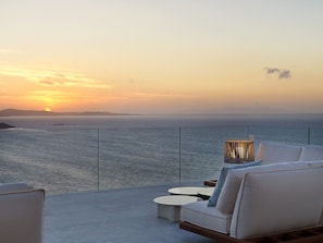 A balcony with a sofa and a table, overlooking a body of water during sunset. - Deos Mykonos (Mykonos)
