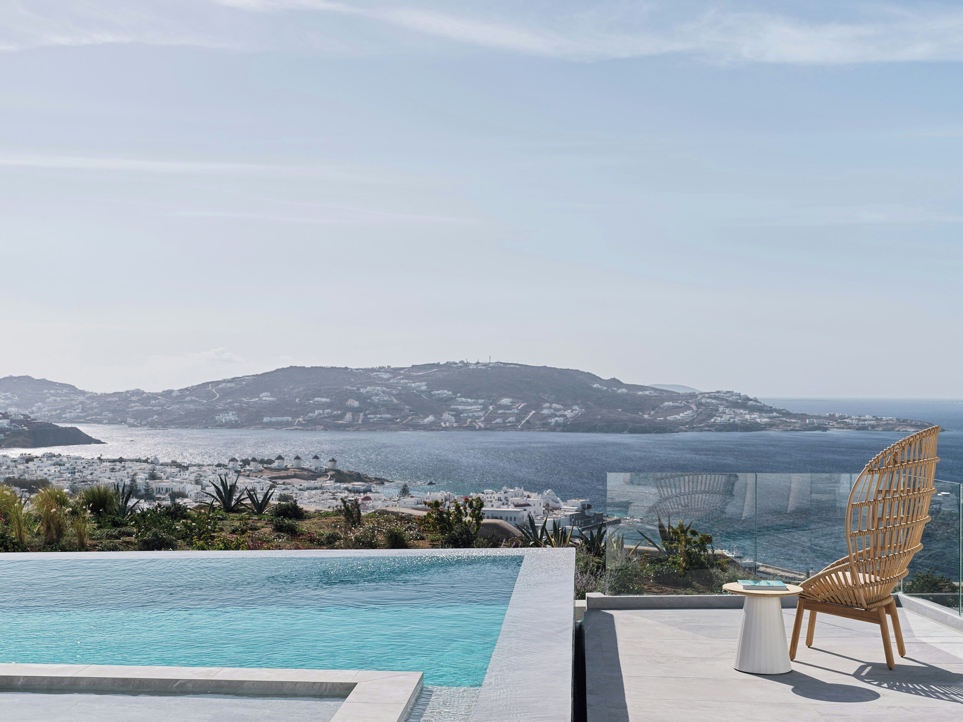 A pool with a view of a coastal town, a wicker chair, and a table.