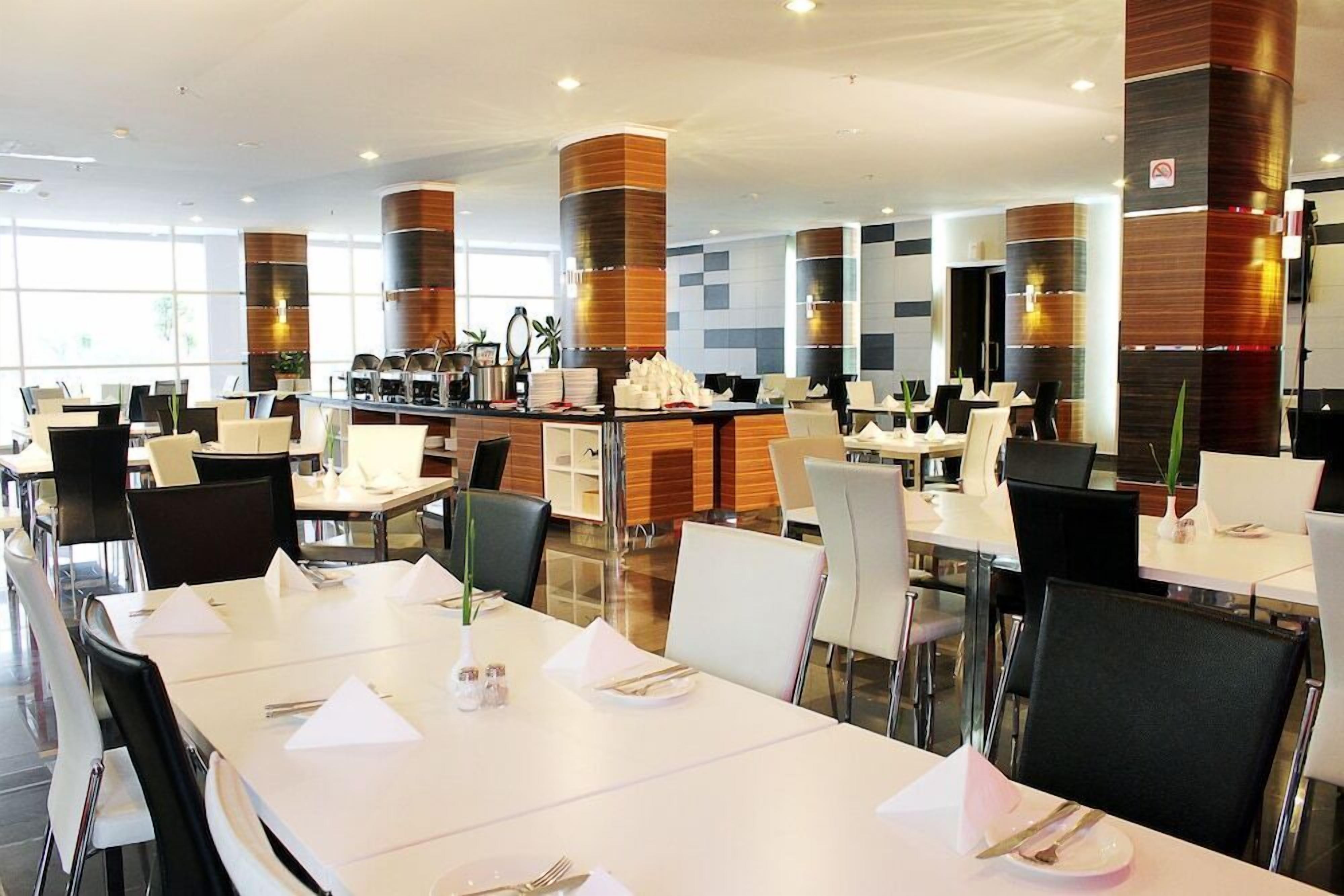 Ein moderner Essbereich mit weißen Tischen, schwarzen Stühlen und einer zentralen Theke mit einer Kaffeemaschine.