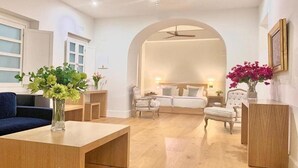 A modern living room with a sofa, a coffee table, and a dining table. There is a bed with a headboard and two chairs. The room has wooden flooring and white walls. - Palacio de la Corredera (Jerez de la Frontera)