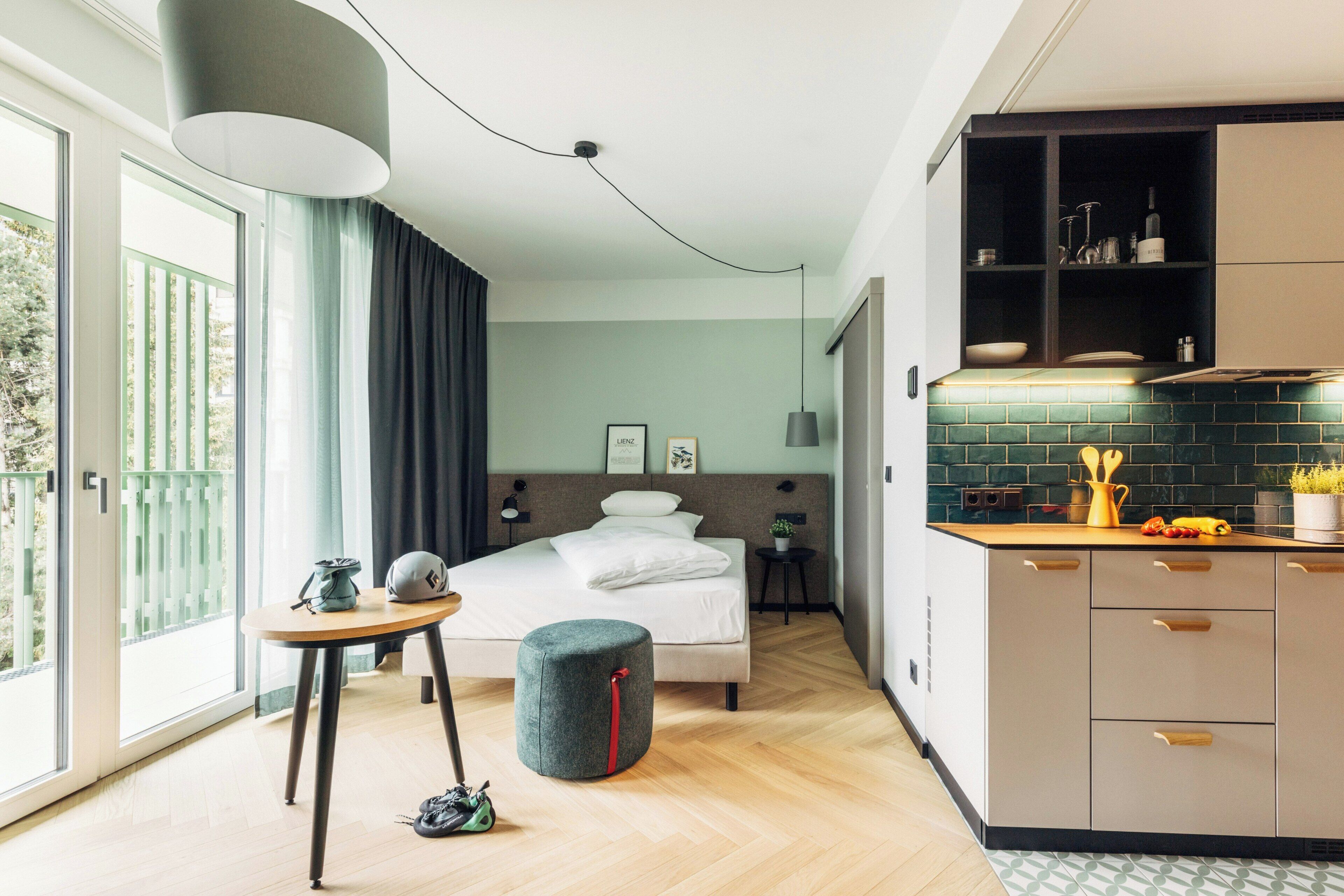 A modern hotel room with a bed, a small table, a stool, a kitchen area with cabinets, and a large window with curtains.