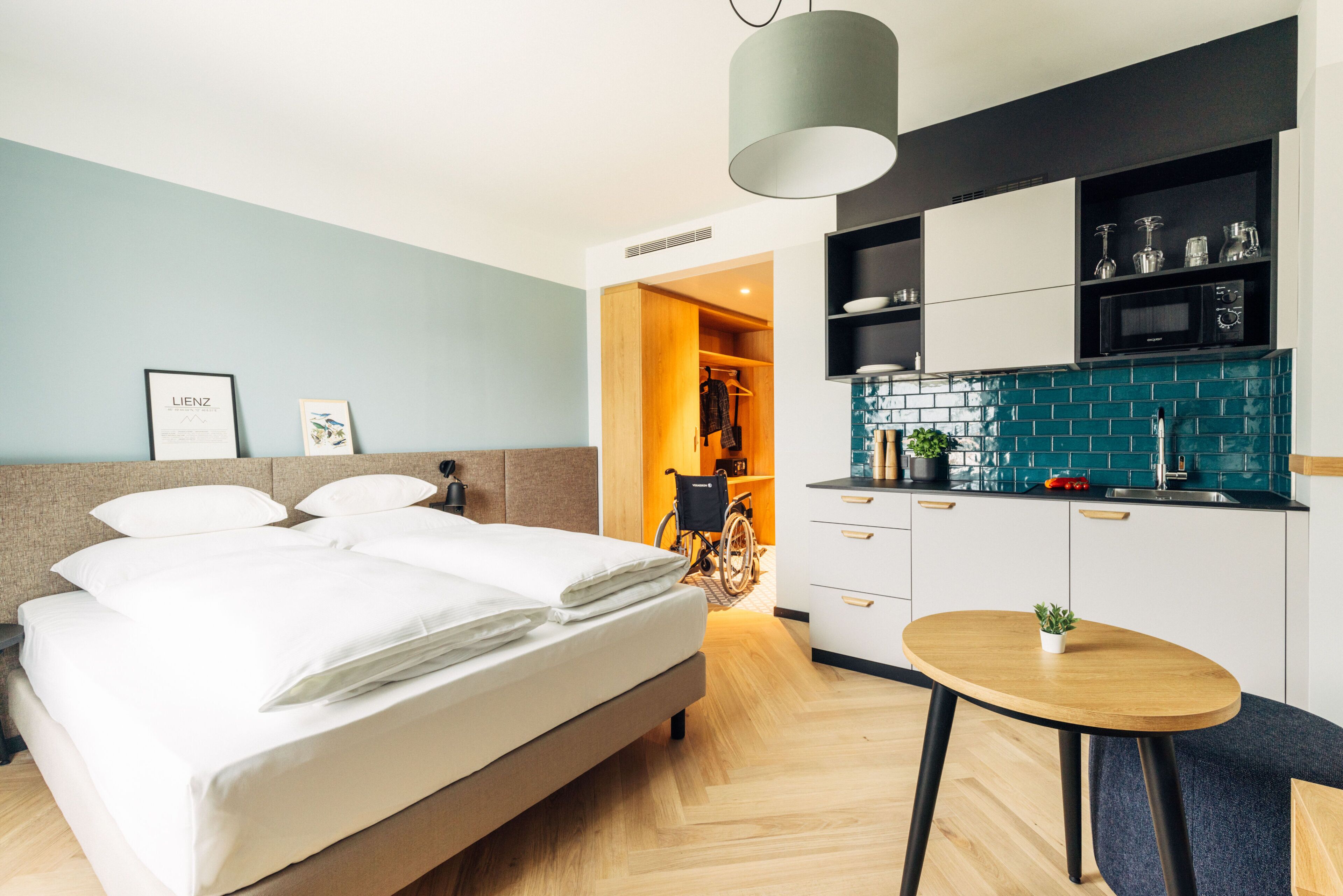 Et moderne hotelværelse med en seng, et lille rundt bord, et tekøkken og en kørestol i baggrunden.