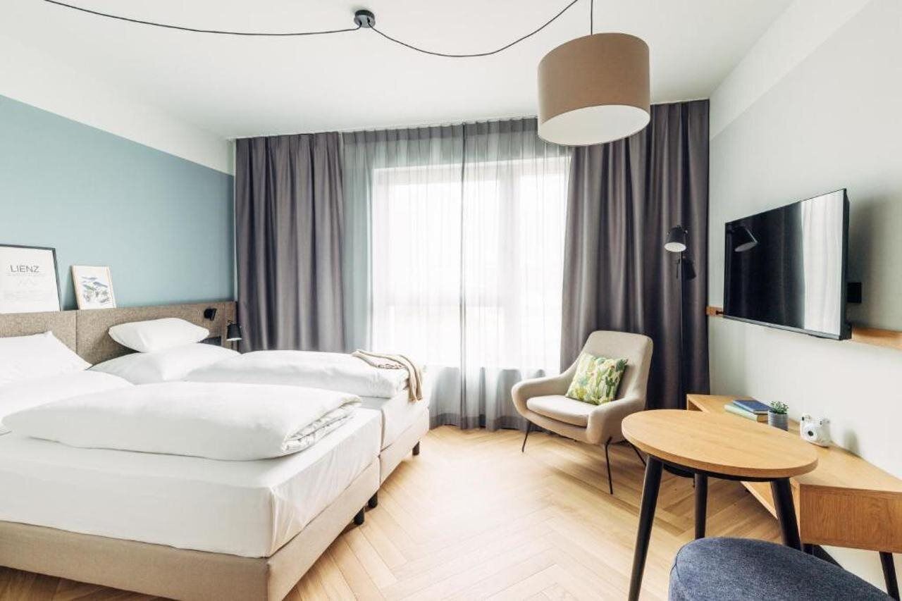 A hotel room with two beds, a desk, a chair, a TV, and a large window with curtains.