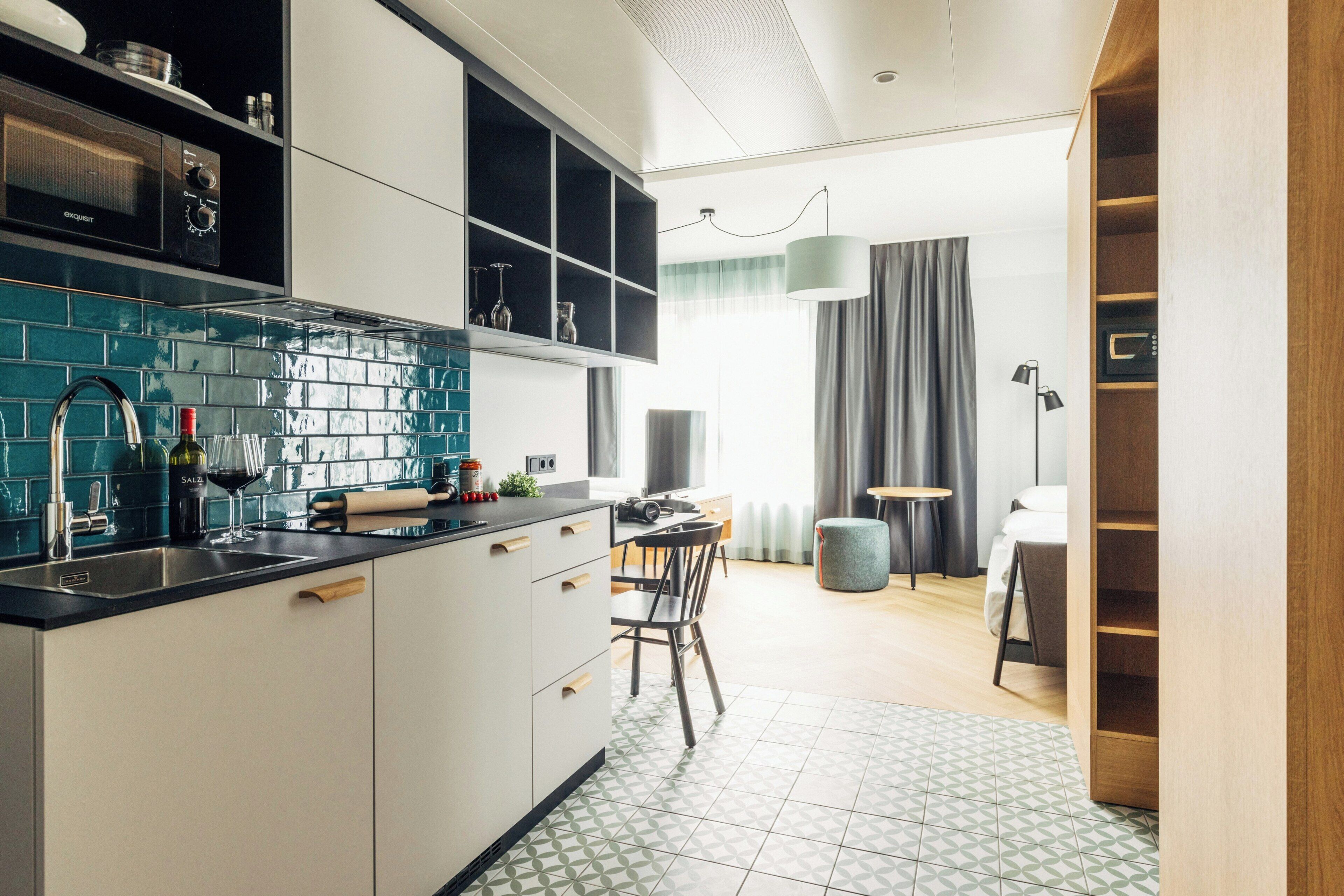 A modern kitchen with a black countertop, white cabinets, and a tiled backsplash. There is a dining area with a table and chairs, a lamp, and a view of a bedroom with a bed and curtains.