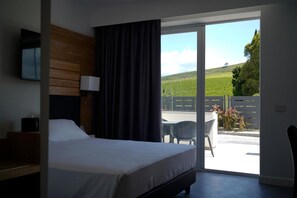 A hotel room with a bed, a desk, a chair, a lamp, and a view of a vineyard through a sliding glass door. - Pm Hotel (Catanzaro)