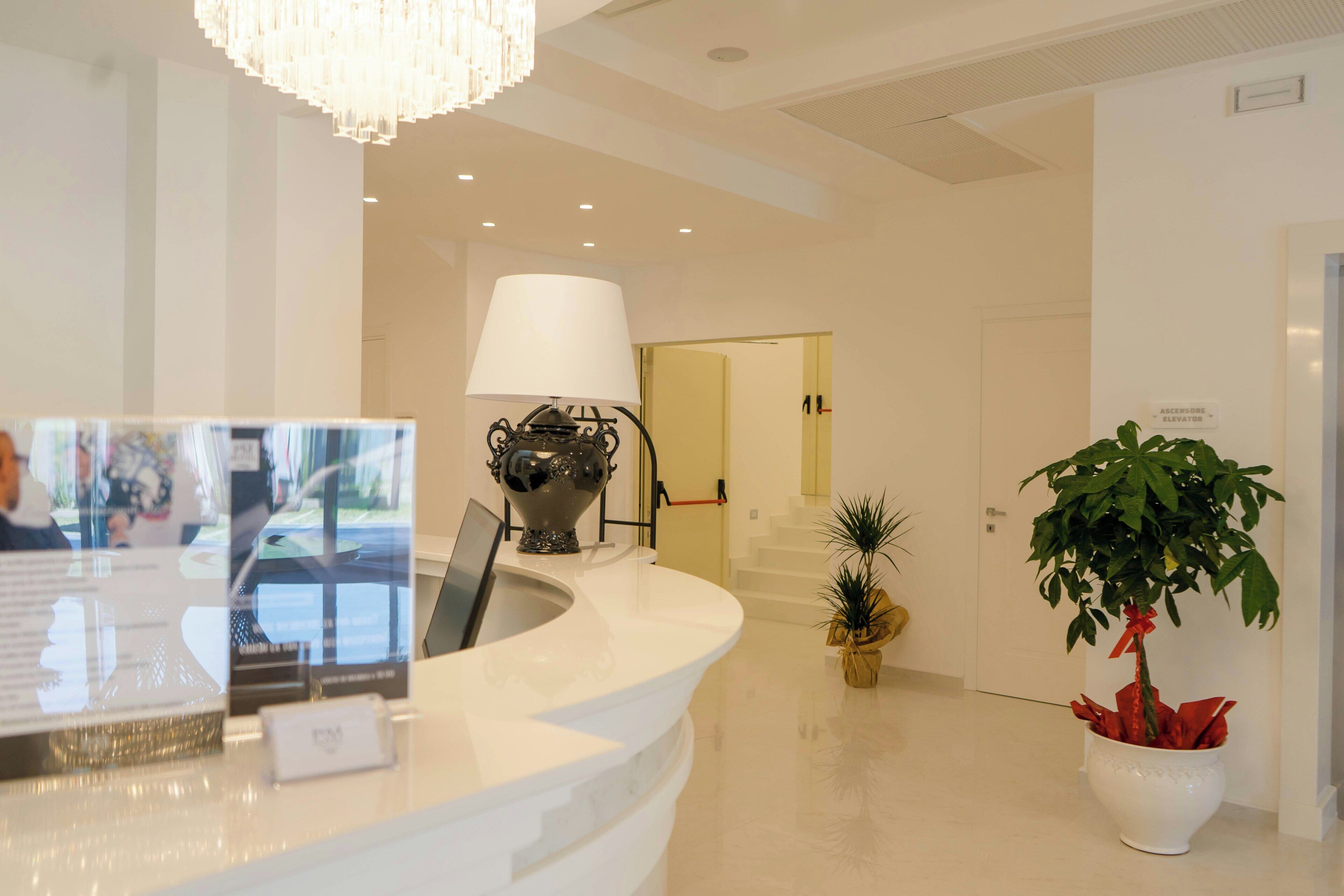 Uma área de recepção de hotel com um balcão branco, um candelabro, um abajur, uma planta em vaso e uma placa indicando um elevador.