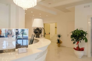 A hotel reception area with a white reception desk, a chandelier, a lamp, a potted plant, and a sign indicating an elevator. - Pm Hotel (Catanzaro)