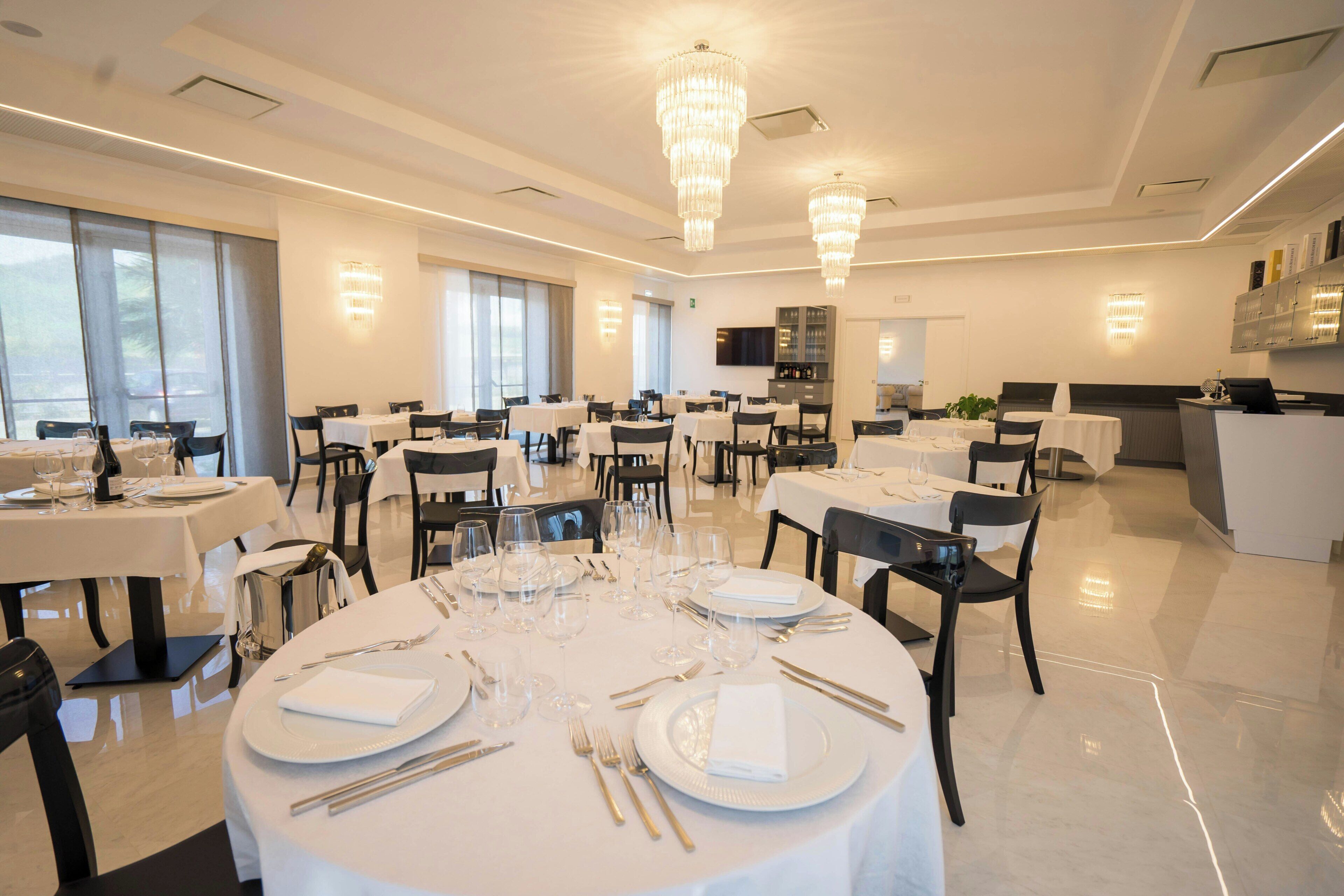 A spacious dining area with multiple tables set for dining, large windows allowing natural light, and modern chandeliers.