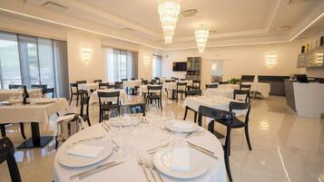 A spacious dining area with multiple tables set for dining, large windows allowing natural light, and modern chandeliers.