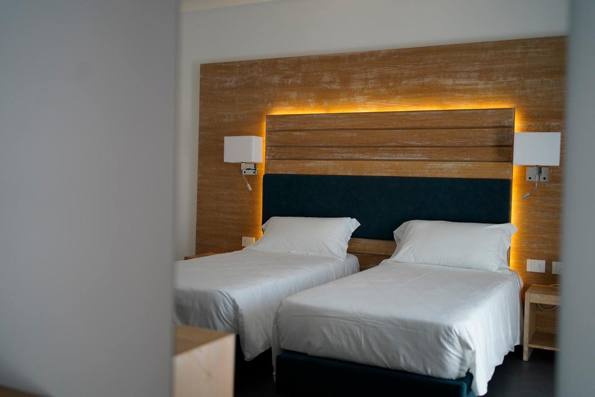 A hotel room with two single beds, wooden headboard, bedside lamps, and a dark-colored headboard.